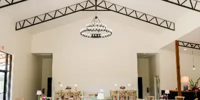 Interior view of a modern barn wedding venue with high ceilings, exposed black trusses, large circular chandeliers, and round tables set with mint green tablecloths.