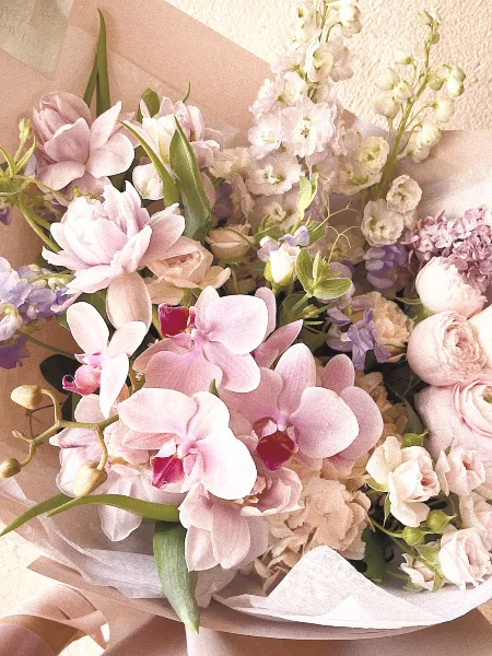 A close-up of a floral arrangement containing pink orchids, roses, and delphiniums wrapped in paper.