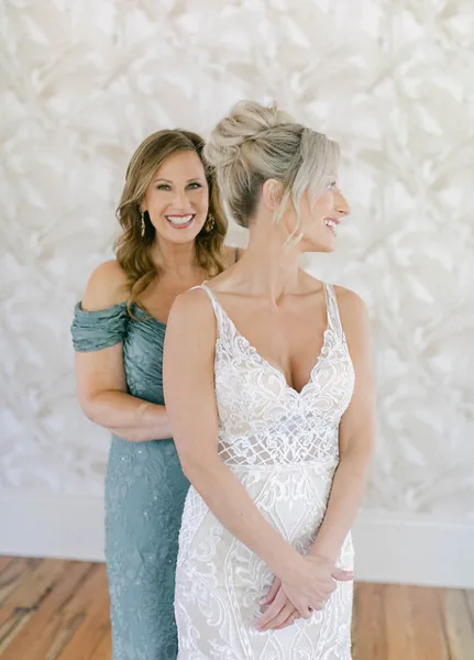 A bride in a white lace gown standing with a smiling woman in a teal dress against a white textured backdrop.