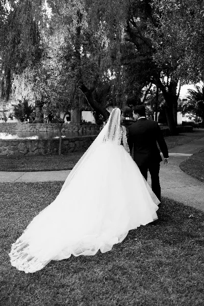 A black-and-white photo of a bride in a long gown and a groom in a dark suit walking away from the camera on a grassy path near a pond.