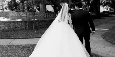 A black-and-white photo of a bride in a long gown and a groom in a dark suit walking away from the camera on a grassy path near a pond.