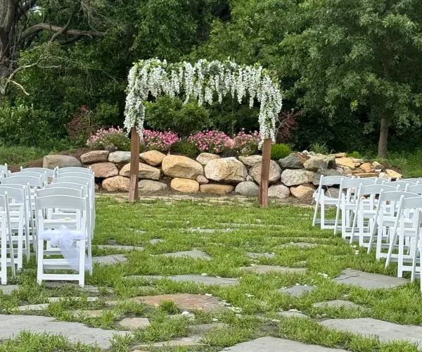 Outdoor wedding ceremony setup with white chairs, a floral arbor, and a rock wall backdrop.