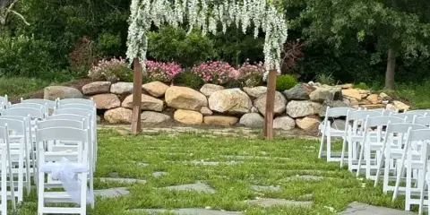 Outdoor wedding ceremony setup with white chairs, a floral arbor, and a rock wall backdrop.