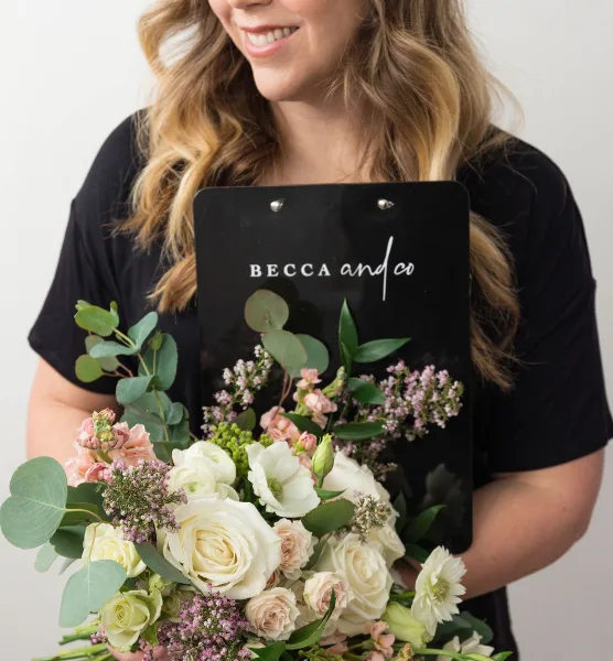 A person holding a wedding bouquet and a black clipboard with the 'Becca & Co.' logo.