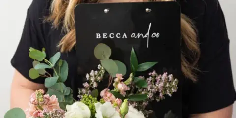 A person holding a wedding bouquet and a black clipboard with the 'Becca & Co.' logo.