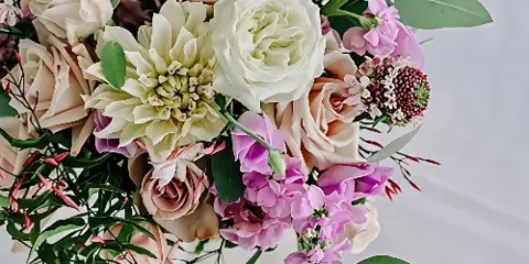 A close-up of a lush floral arrangement featuring white and blush roses, a large cream dahlia, and pink lisianthus.