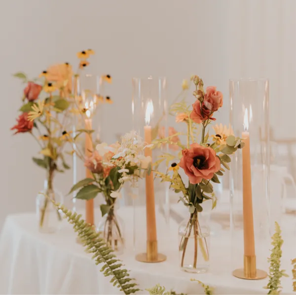 A wedding table centerpiece featuring peach roses, yellow wildflowers, and greenery in small glass vases alongside tall taper candles.
