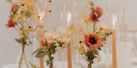 A wedding table centerpiece featuring peach roses, yellow wildflowers, and greenery in small glass vases alongside tall taper candles.