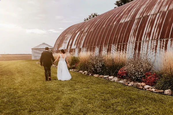 A bride and groom walking hand-in-hand on a grassy lawn next to a large, curved metal building with a garden bed.