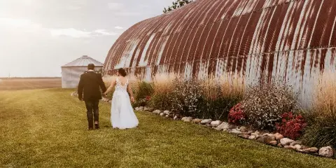 A bride and groom walking hand-in-hand on a grassy lawn next to a large, curved metal building with a garden bed.
