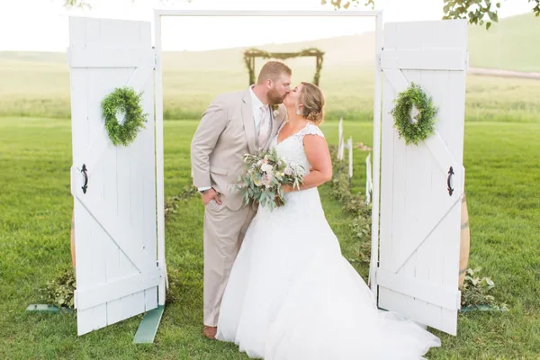 A bride and groom kissing under a white wooden archway decorated with greenery wreaths in a grassy field.