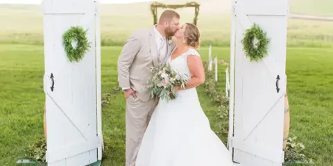 A bride and groom kissing under a white wooden archway decorated with greenery wreaths in a grassy field.