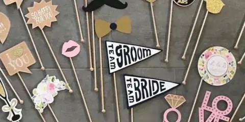 An assortment of wedding-themed photo booth props including flags, glasses, and mustaches arranged on a grey surface.
