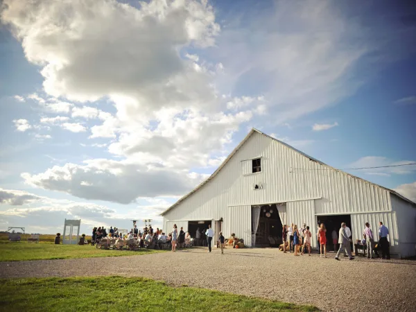 A white rustic barn venue with an outdoor wedding reception and guests gathered on the grounds under a blue sky.