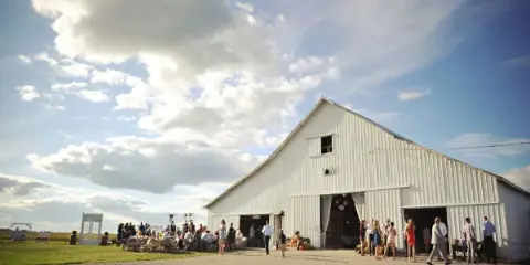 A white rustic barn venue with an outdoor wedding reception and guests gathered on the grounds under a blue sky.