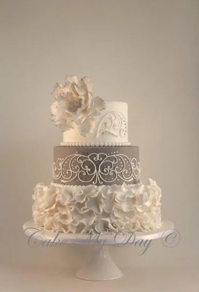 A three-tiered wedding cake featuring white ruffles, a grey tier with white scrollwork, and a large white sugar flower on top.