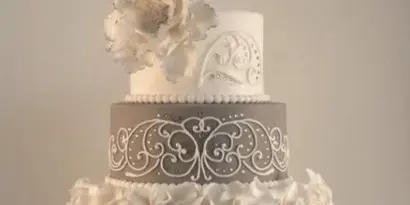 A three-tiered wedding cake featuring white ruffles, a grey tier with white scrollwork, and a large white sugar flower on top.