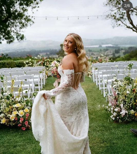 A smiling bride with styled blonde wavy hair and a lace wedding dress standing in an outdoor ceremony aisle.