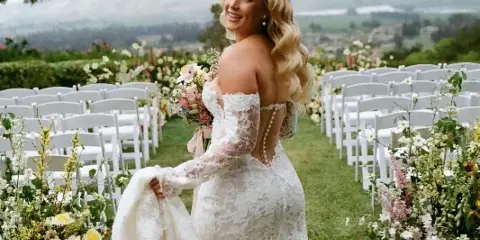 A smiling bride with styled blonde wavy hair and a lace wedding dress standing in an outdoor ceremony aisle.