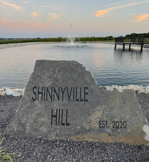 A large stone sign reading 'Shinnyville Hill Est. 2020' in the foreground with a pond, fountain, and wooden dock in the background at sunset.