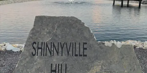A large stone sign reading 'Shinnyville Hill Est. 2020' in the foreground with a pond, fountain, and wooden dock in the background at sunset.