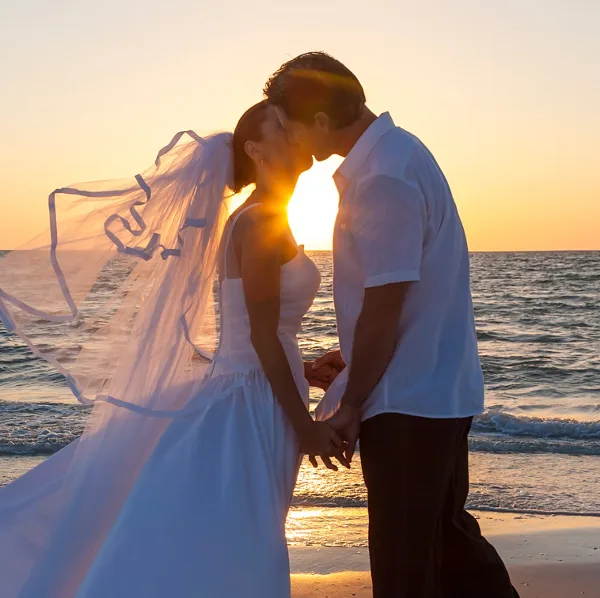 A bride and groom kissing on a sandy beach with the sun setting over the ocean in the background.