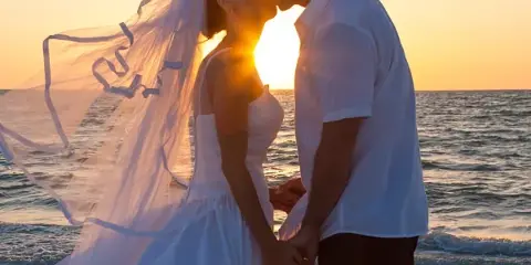 A bride and groom kissing on a sandy beach with the sun setting over the ocean in the background.