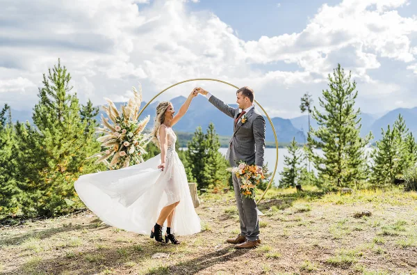 A bride and groom dancing under a gold arch decorated with a large floral arrangement and pampas grass in an outdoor mountain setting.