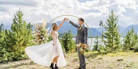 A bride and groom dancing under a gold arch decorated with a large floral arrangement and pampas grass in an outdoor mountain setting.