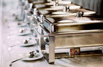 A row of stainless steel chafing dishes set up for a buffet with serving spoons on a table.