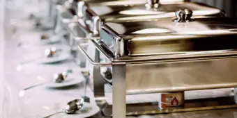 A row of stainless steel chafing dishes set up for a buffet with serving spoons on a table.