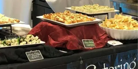 A catering buffet table displaying various gourmet appetizers, including olive feta and potato artichoke bites, with a 'Catering' banner.
