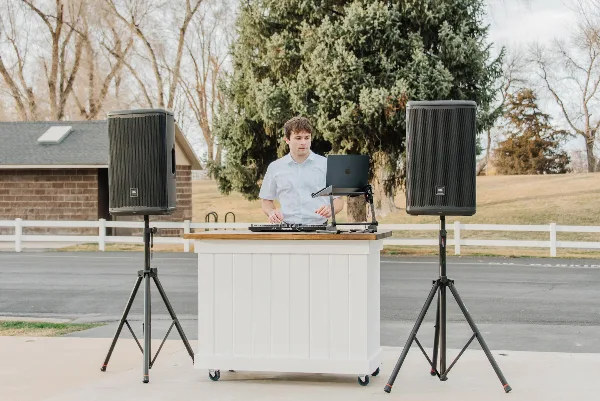 A DJ operating a laptop and mixer at a white podium, flanked by two large speakers on stands, in an outdoor setting.