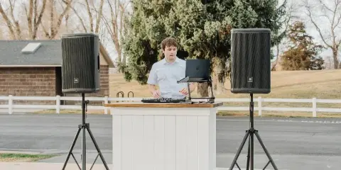A DJ operating a laptop and mixer at a white podium, flanked by two large speakers on stands, in an outdoor setting.