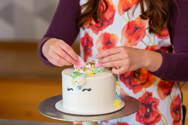 A person wearing a red poppy apron decorates a white frosted cake with sprinkles and fondant toppers on a metal cake stand.