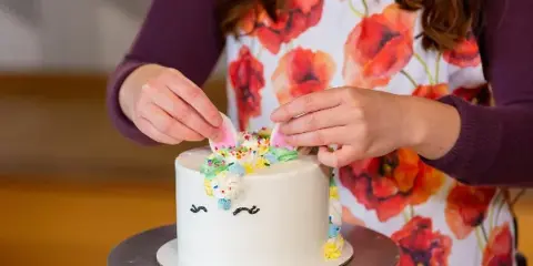 A person wearing a red poppy apron decorates a white frosted cake with sprinkles and fondant toppers on a metal cake stand.