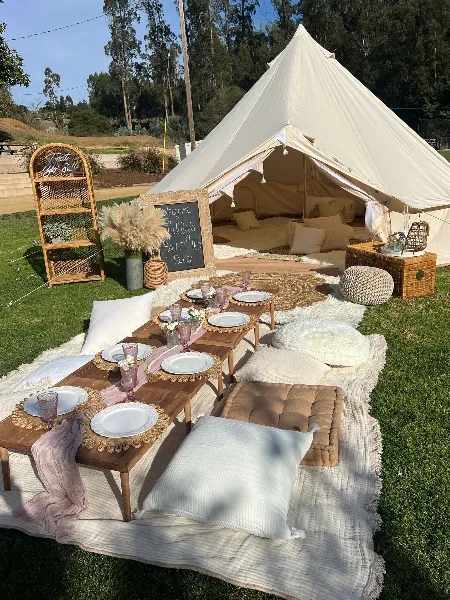 A styled outdoor event setup featuring a white bell tent, lounge seating, and a boho picnic arrangement on a grassy lawn.