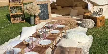 A styled outdoor event setup featuring a white bell tent, lounge seating, and a boho picnic arrangement on a grassy lawn.