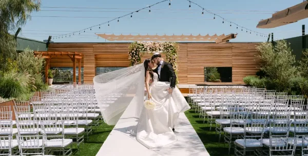 A bride and groom sharing a kiss at an outdoor wedding ceremony with white chairs and a floral arch.