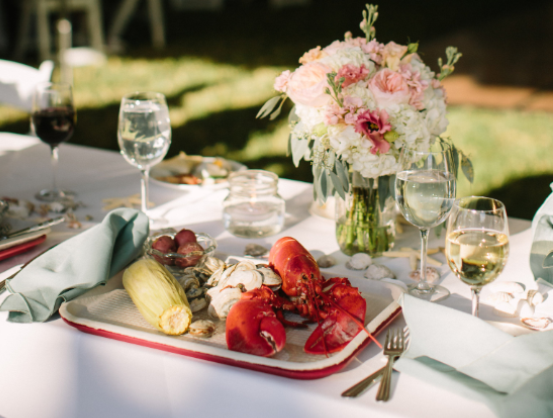 A catering table set with a platter of lobster, corn, and clams, accompanied by wine glasses and a floral centerpiece.