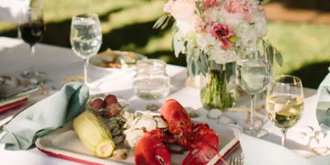 A catering table set with a platter of lobster, corn, and clams, accompanied by wine glasses and a floral centerpiece.