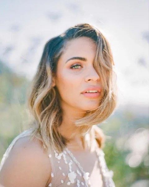 A professional portrait of a woman with styled wavy hair and makeup, wearing a white beaded top.