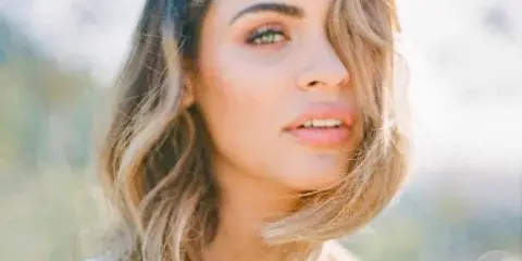 A professional portrait of a woman with styled wavy hair and makeup, wearing a white beaded top.