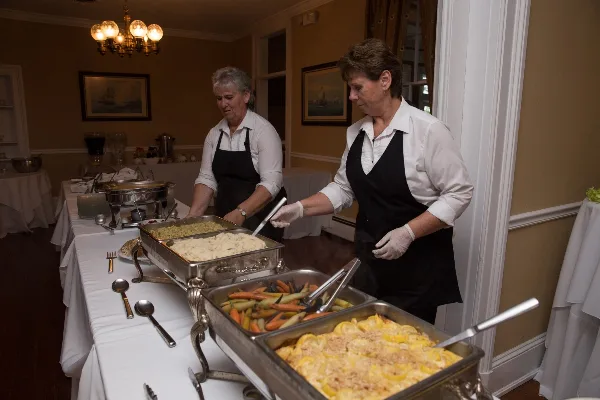 Two catering staff members in white shirts and black aprons serving food from chafing dishes at an event.