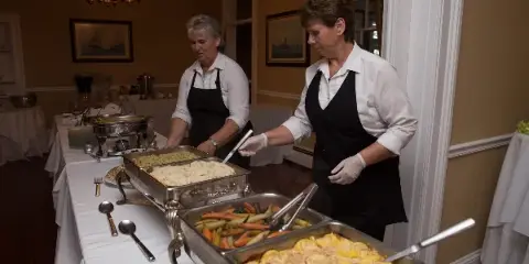 Two catering staff members in white shirts and black aprons serving food from chafing dishes at an event.