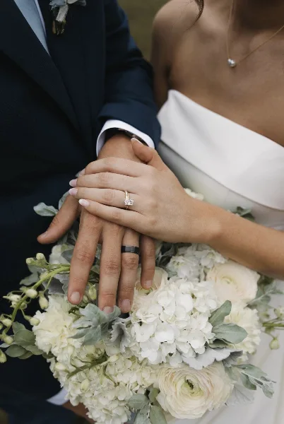Close-up of a bride and groom's hands holding a white wedding bouquet with hydrangeas and eucalyptus.