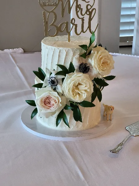 A white two-tier wedding cake decorated with white roses and greenery, featuring a gold 'Mrs Baril' topper.