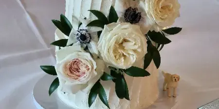 A white two-tier wedding cake decorated with white roses and greenery, featuring a gold 'Mrs Baril' topper.