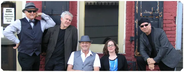 Group photo of the Shaky Ground Band members standing and sitting in front of a red brick building.