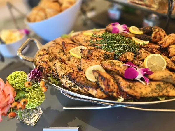 A large silver platter filled with grilled chicken pieces garnished with lemon slices, fresh herbs, and orchids, set on a table with a floral arrangement.